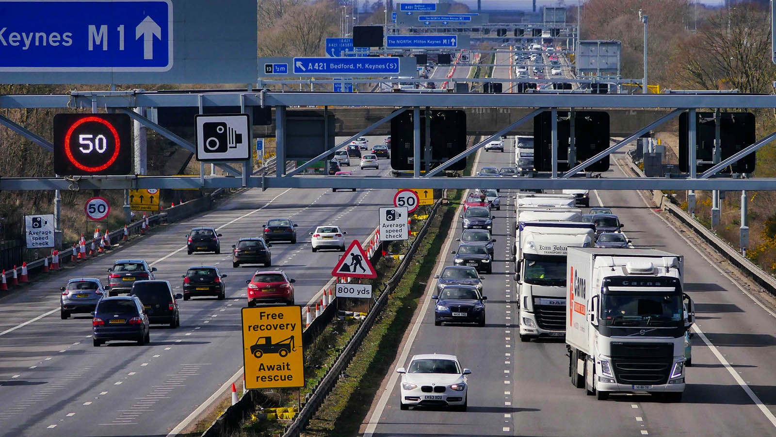 A UK motorway with lots of traffic, an overhead gantry with live speed limits and an average speed check zone